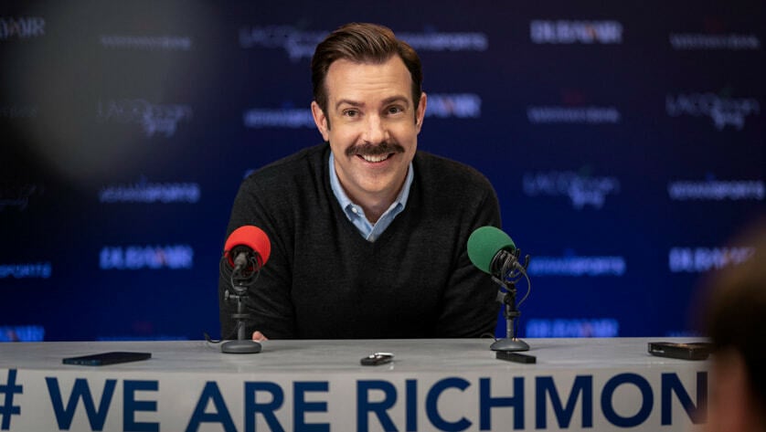 A smiling man with a mustache sits behind two colorful microphones.
