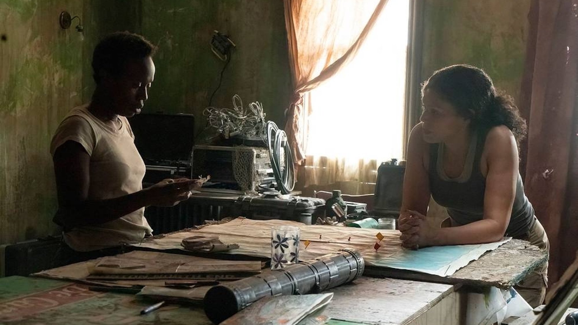 Two women wearing military tank tops stand considering a map on a table in a dilapidated apartment.