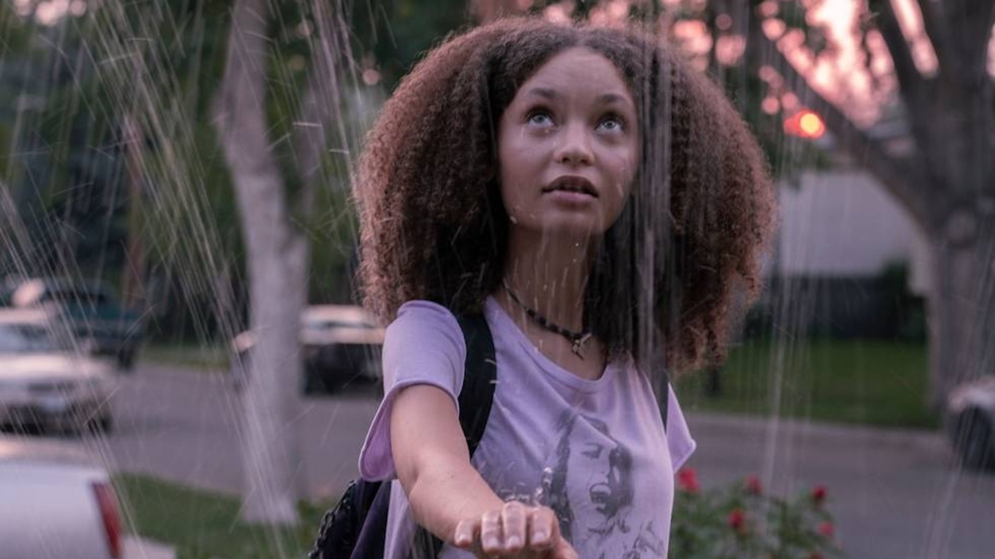 A teen girl holds her hand in a suburban sprinkler.